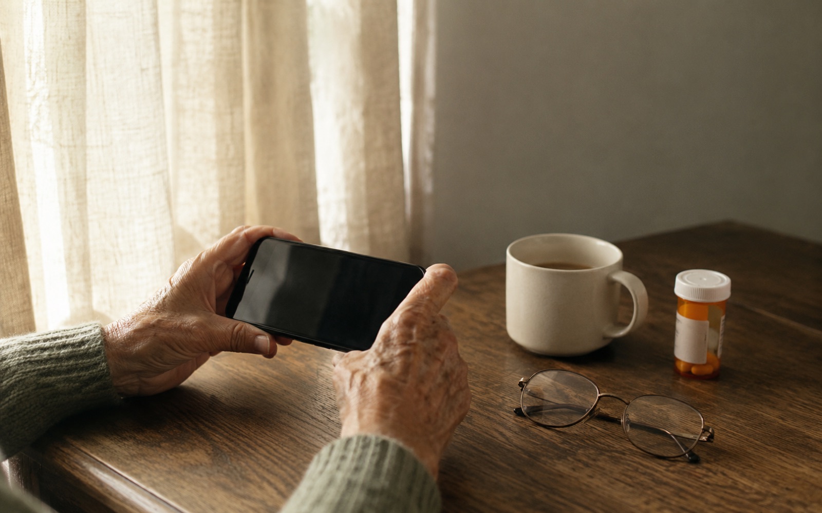 Morning light on a worn oak table: a senior's hands holding a smartphone next to a coffee cup and an amber prescription bottle.